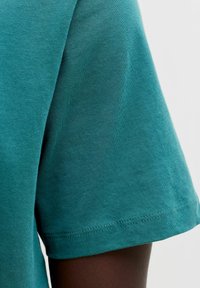 Tissu en coton bleu sarcelle à manches courtes sur un bras à peau foncée, gros plan montrant le détail des coutures sur l'ourlet de la manche.