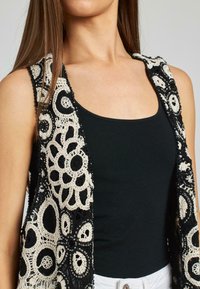 Schwarze und cremefarbene gehäkelte Weste mit komplizierten Blumenmustern, offenem Vorderteil und mit Perlen verzierten Akzenten, getragen mit einem schwarzen Tanktop.