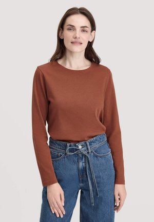 Langärmliges Oberteil mit Rundhalsausschnitt aus rostbraunem Stoff, kombiniert mit hoch taillierten blauen Jeans aus Denim, die über eine gebundene Taille und seitliche Taschen verfügen.