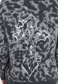 Grijze gebreide trui met een groot wit geborduurd kruisontwerp op de achterkant, voorzien van bloemaccenten en zwevende lijnen.