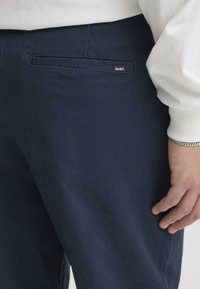 Teget hlače od pamuka glatke teksture. Imaju stražnji džep i malu oznaku brenda na lijevoj strani.