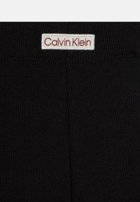 Fekete bordázott anyagú derékszíj, fehér címkével, rajta piros szöveggel: "Calvin Klein". A kialakítás egyszerű és minimalista.
