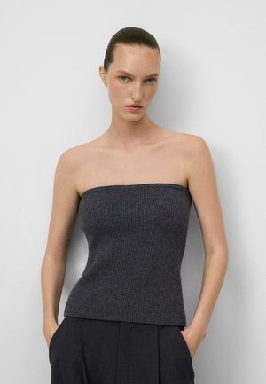 Vrouw met strak naar achter gekamd haar, gekleed in een donkergrijze strapless ribbeltop en zwarte broek met haar handen in de zakken, staande tegen een witte muur.