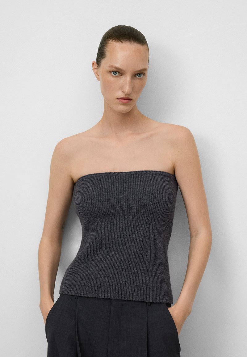 Femme aux cheveux coiffés en arrière, portant un haut sans manches à rayures gris foncé et un pantalon noir, les mains dans les poches, adossée à un mur blanc.