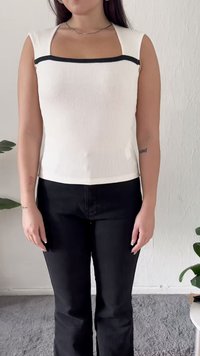 Woman wearing a white sleeveless ribbed top with black trim and black pants, standing indoors against a white textured wall.