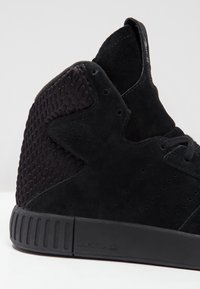 Černé vysoké tenisky s semišovými a síťovinovými panely, strukturovanými boky, kulatými tkaničkami a gumovou podrážkou se vodorovnými drážkami.