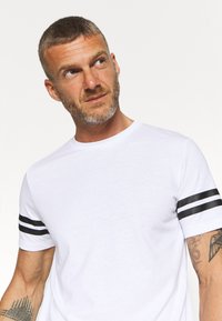 T-shirt bianco in cotone con dettagli a strisce nere sulle maniche corte, scollo rotondo e una texture liscia. Presenta una vestibilità casual e un design minimalista.