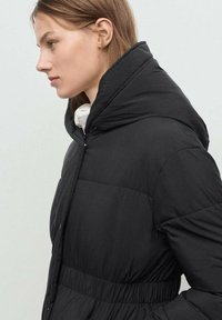 Mujer joven vestida con una chaqueta acolchada negra de cuello alto, mirando hacia la izquierda contra un fondo claro y liso.