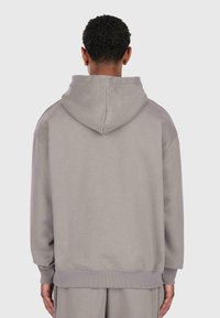 Uomo con capelli ricci corti indossa un grande hoodie grigio chiaro e pantaloni abbinati, visto di spalle su uno sfondo semplice.