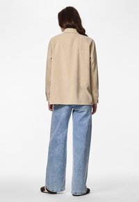 Beigevärinen ylihuppuinen puuvillakorduroy-paita pitkähihainen takakappaleella, yhdistetty vaaleansinisien leveälahkeisten farkkujen kanssa. Yksinkertaiset ruskeat jalkineet.