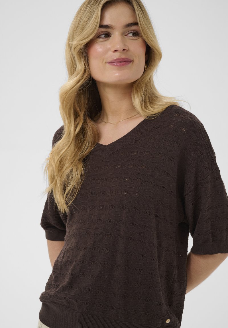 Femme blonde portant un pull à manches courtes texturé brun foncé à col en V, regardant légèrement sur le côté, souriant doucement.