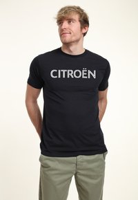 Černé tričko s krátkým rukávem s nápisem "CITROËN" bílým písmem přes hruď. Vyrobeno z hladkého materiálu. Nosené s světle zelenými kalhotami.