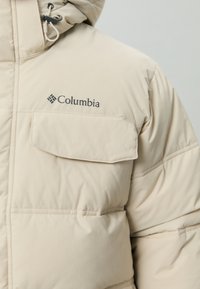Beige pufferjas met een capuchon, met een grote vo zak, gestikte secties en een zwart Columbia-logo op de borst.