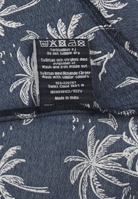Blauw stof met een wit palmenbladpatroon. Verzorgingslabel bevestigd. Materiaal lijkt lichtgewicht, met een zachte textuur en afgewerkte randen.