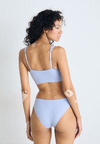 Kvinna med vitiligo på armar och rygg, klädd i en ljusblå ribbad bikinitopp, står mot en enhetlig ljus bakgrund och ser över sin axel.