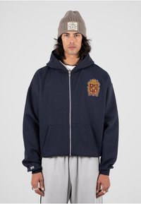 Sudadera con cremallera de color azul marino con un gran escudo bordado en el pecho, bolsillos frontales y capucha con cordón. Lleva debajo una camisa blanca y pantalones grises.