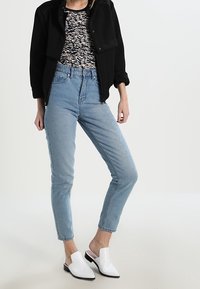 Chaqueta negra corta, blusa con estampado, vaqueros de tiro alto azul claro y mocasines blancos con un pequeño tacón. Líneas limpias y diseño moderno.