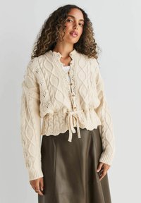 Creme kabelstrik cardigan med en cropped design, der har et bånd i taljen, knaplukning og ribbede manchetter. Bæres over en mørk læderskirt.