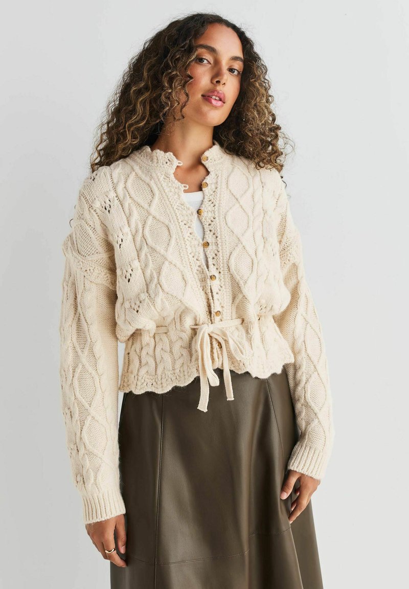 Creme kabelstrik cardigan med en cropped design, der har et bånd i taljen, knaplukning og ribbede manchetter. Bæres over en mørk læderskirt.
