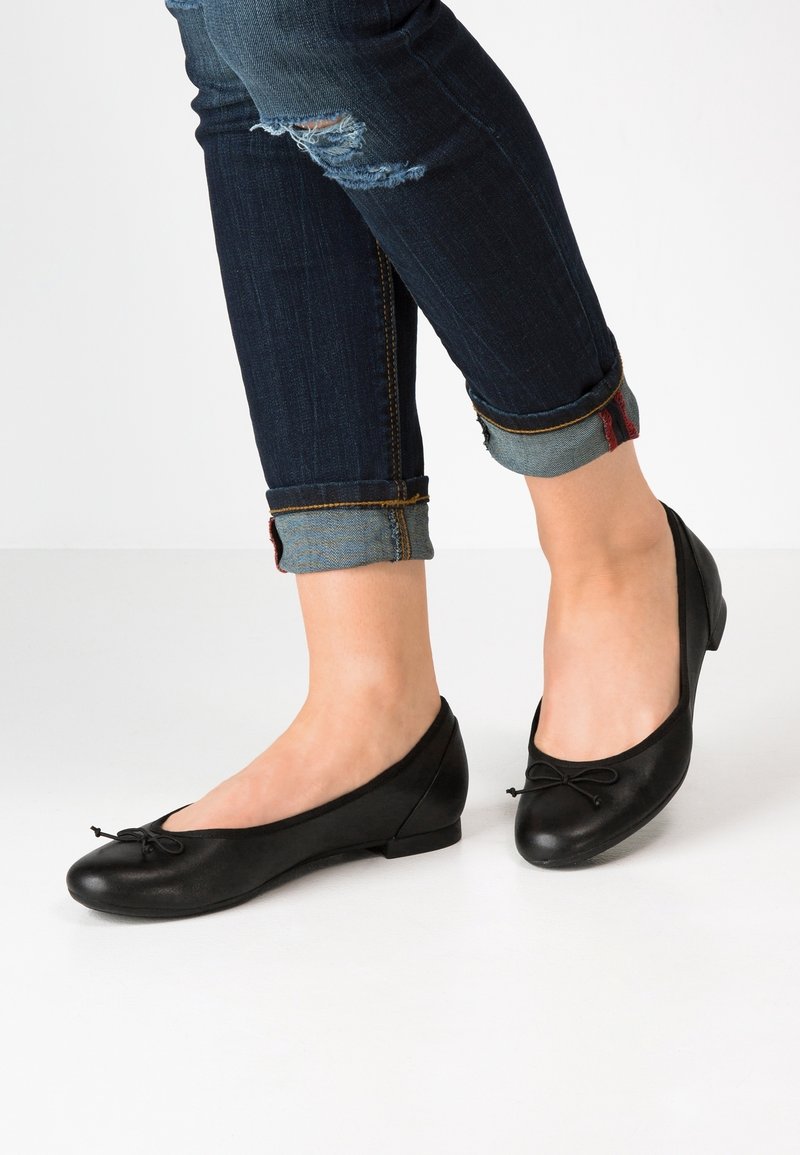 Jambes portant un jean foncé retroussé et déchiré ainsi que des ballerines noires avec de petits nœuds décoratifs sur un fond blanc.