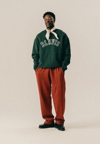 Mann steht mit den Händen in den Taschen, trägt einen grünen BLKVIS Sweatshirt, rote Hose, schwarze Schuhe, Brille und ein weißes Bandana um den Hals.