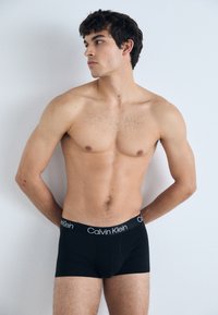 Schwarze Baumwoll-Boxershorts mit einem weißen "Calvin Klein"-Logo am Bund, präsentiert an einem männlichen Model vor einem schlichten Hintergrund.