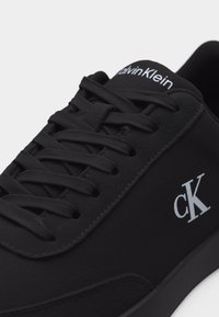 Baskets Calvin Klein noires avec lacets noirs, logo "cK" blanc sur le côté, et le texte "Calvin Klein" sur la languette, sur un fond uni.