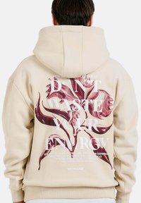 Beige huvtröja med en stor blommig grafik och textdesign i mörkröd och vit på baksidan. Mjukt bomullsmaterial.