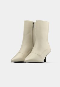 Paire de bottines beiges mi-mollet à bout pointu, avec fermeture éclair latérale et petits talons stilettos sur fond clair uni.