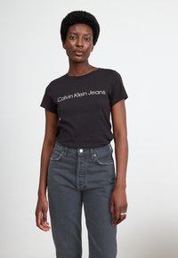 Černé bavlněné tričko s bílým logem "Calvin Klein Jeans", spárované s šedými džíny s vysokým pasem, které mají přední kapsy a zip.
