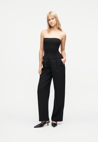 Γυναίκα με ξανθά μαλλιά που φοράει μαύρο τοπ χωρίς τιράντες, μαύρο παντελόνι με φαρδιά μπατζάκια και ψηλή μέση, και μαύρες γόβες με μυτερή μύτη, στέκεται με το ένα χέρι στην τσέπη.