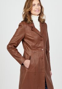Bruin leren jasje met een op maat gemaakt ontwerp, geknipte revers, dubbel knopen en zijzakken. Gladde textuur, lange lengte tot de knieën.