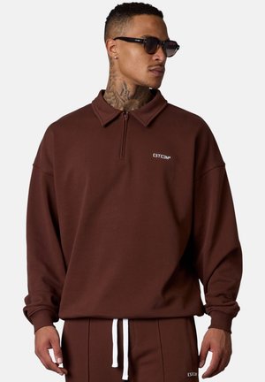Pull-over zippé marron avec col, fabriqué en tissu doux, arborant un petit logo blanc et une coupe décontractée. Associé à un pantalon assorti.