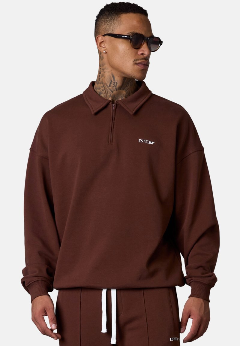Pull-over zippé marron avec col, fabriqué en tissu doux, arborant un petit logo blanc et une coupe décontractée. Associé à un pantalon assorti.