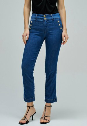 Mujer con vaqueros azules de talle alto con botones dorados en los bolsillos y la cintura, combinados con sandalias negras de tacón con tiras.