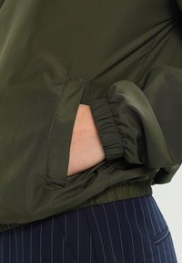 Hånd i lommen på olivengrønn jakke med elastisk mansjett, kombinert med marineblå bukser med småstriper.