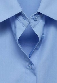 Chemise bleu clair à col, vue de face, avec deux boutons fermés et un déboutonné, montrant la couche intérieure du tissu.