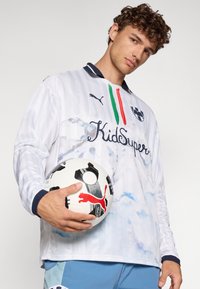 Weißes Fußballtrikot mit langen Ärmeln und vertikalen Streifen, blauer Kragen und "KidSuper"-Logo; hält einen gemusterten Fußball.