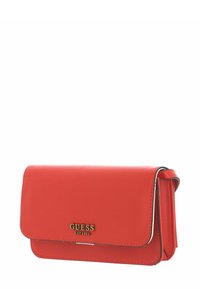 Sac bandoulière en simili cuir rouge avec ferrures dorées, fermeture à rabat et logo "GUESS EST 1981" sur le devant. Forme compacte et rectangulaire.