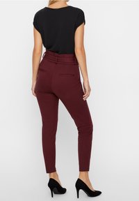 Pantalon ajusté bordeaux avec une taille ceinturée, texture lisse, poches arrière, associé à un haut noir à manches courtes et des talons hauts noirs.