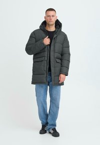 Dunjakke i mørkegrå med hætte, præget af et quiltet design, frontlynlås, to sidelommer, båret over en sort top og blå jeans.