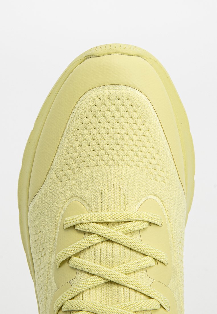 Zapatilla deportiva amarilla con parte superior de tejido texturizado, puntera lisa y cordones planos. Presenta una suela redondeada y detalles perforados.