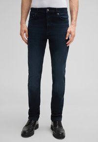 Ciemnoniebieskie dżinsy o slim fit, z pięcioma kieszeniami, subtelną teksturą i srebrnym guzikiem w talii. Na nogach noszone są czarne, wiązane buty.