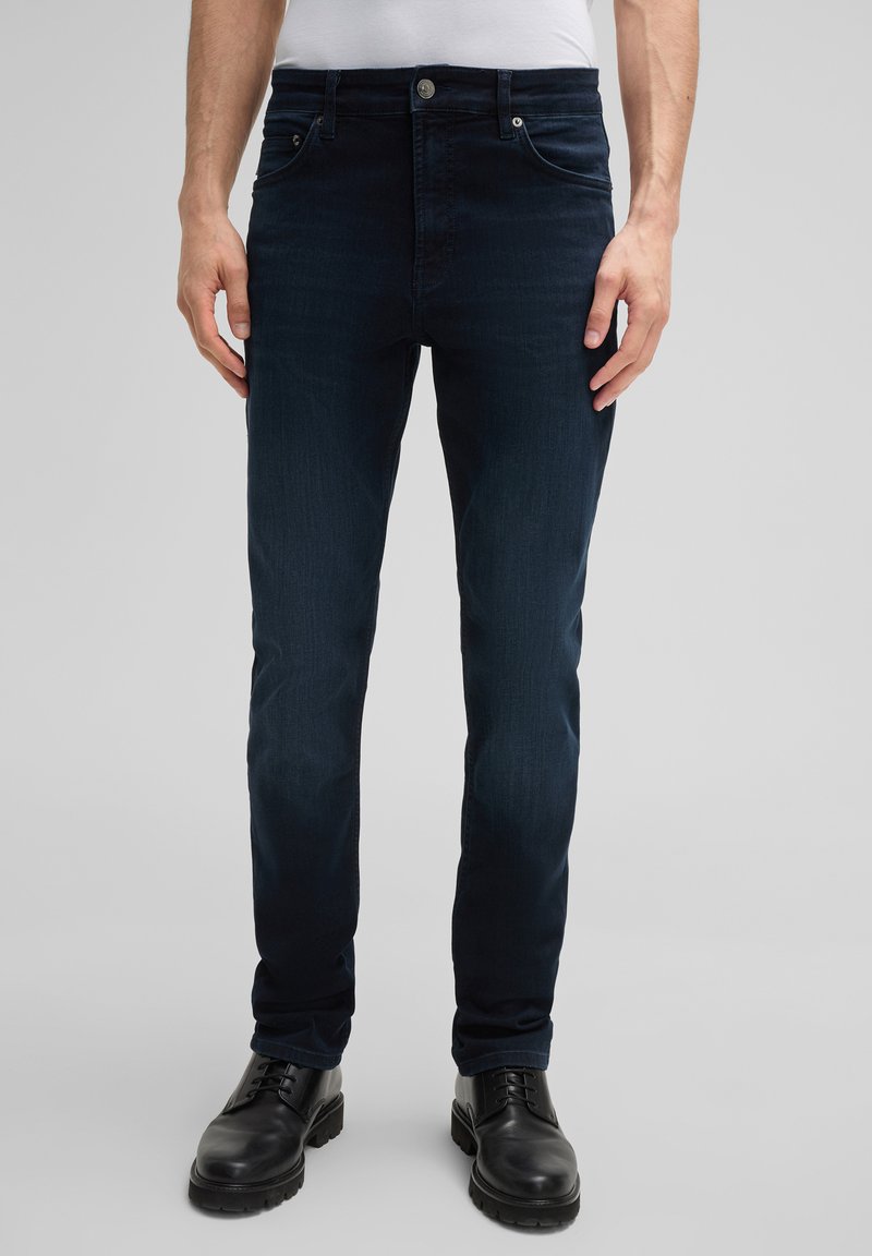 Ciemnoniebieskie dżinsy o slim fit, z pięcioma kieszeniami, subtelną teksturą i srebrnym guzikiem w talii. Na nogach noszone są czarne, wiązane buty.