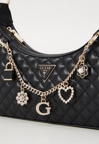 Black quilted handbag with gold hardware. Features a logo plaque and decorative charms, including a flower and heart, on a gold chain.