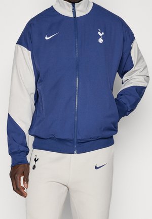 Homme portant une veste zippée bleue et grise et un pantalon gris clair avec les logos Tottenham Hotspur et Nike visibles sur les deux vêtements.