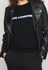 Chaqueta de motocicleta de cuero negro con cremallera plateada, usada sobre una camiseta negra con un logo de texto azul y morado. Pantalones negros completan el look.