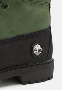 Roheline sametking, musta kummi tallaga, millel on ringikujuline logo puu kujutisega. Nähtavad tugevdõmmed ja nöörimise disainid.