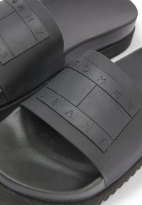 Black slide sandals featuring a textured rubber strap with embossed "TOMMY JEANS" logo, contoured footbed, and durable sole.