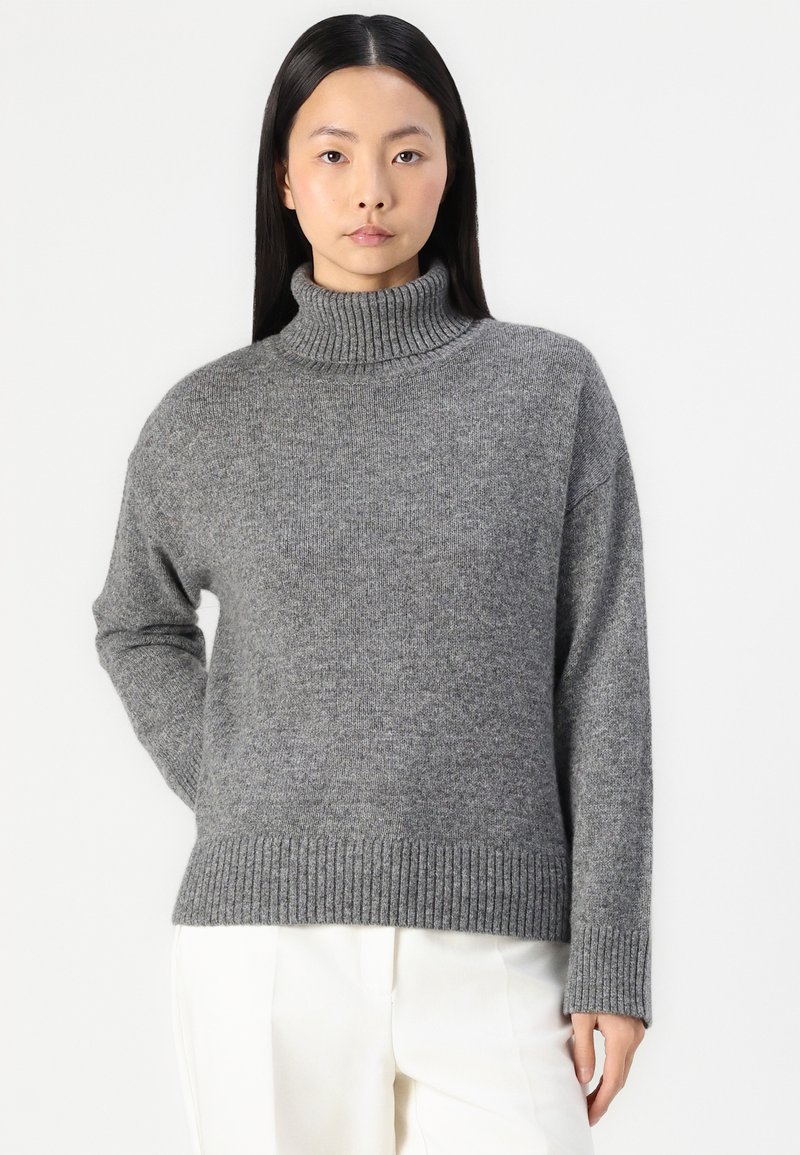 Grauer Rollkragenpullover aus Strickstoff, mit lockerer Passform, gerippten Bündchen und Saum sowie einer weichen Textur. Kombiniert mit hellen Hosen.